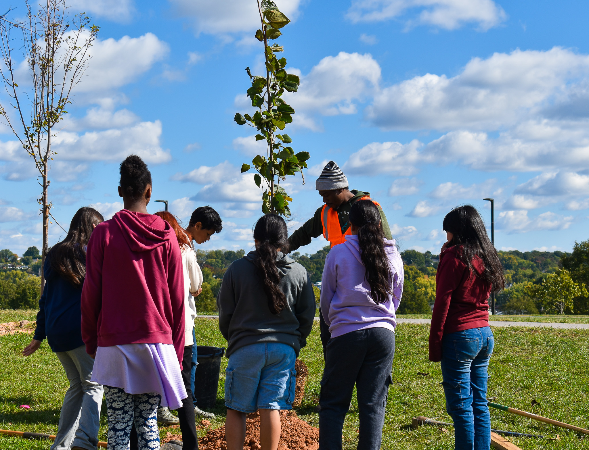 A group of sixth grade students gather around a BTT Neighborhood Forester holding a juvenile tree as he describes the planting process.