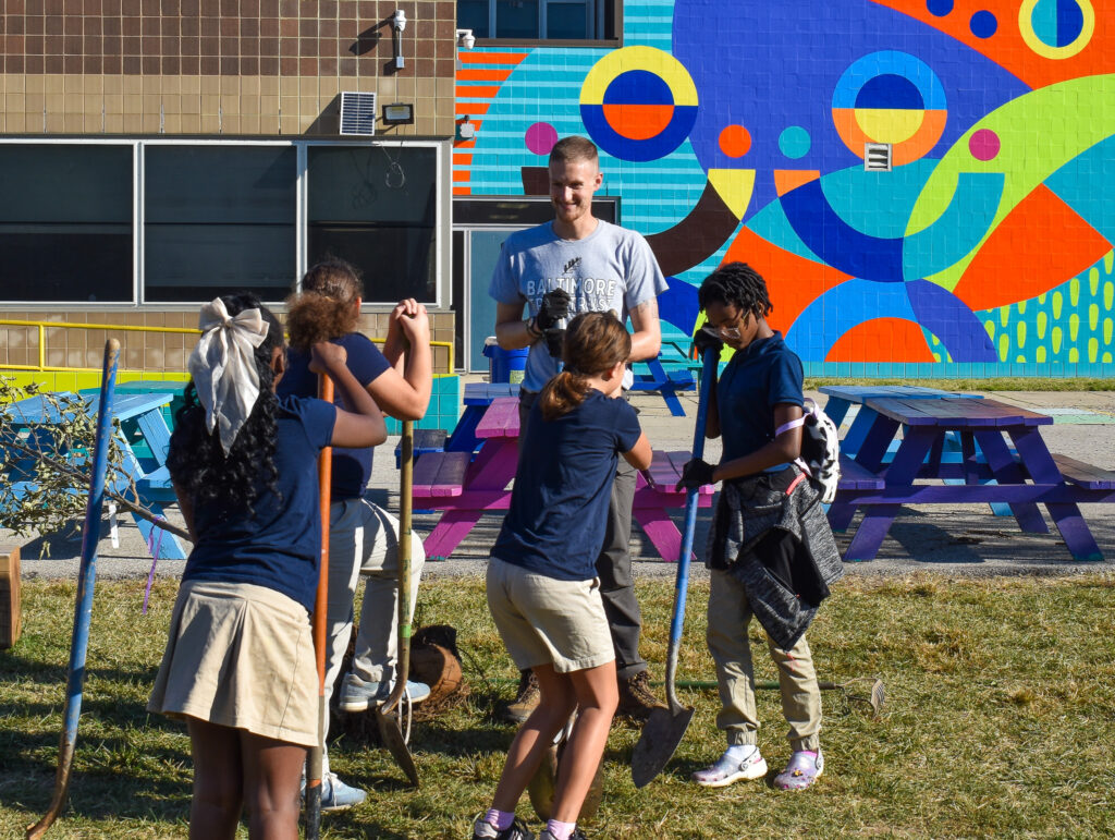 Photo of a male Chesapeake Climate and Conservation Corps member planting trees at a school with a group of four children.
