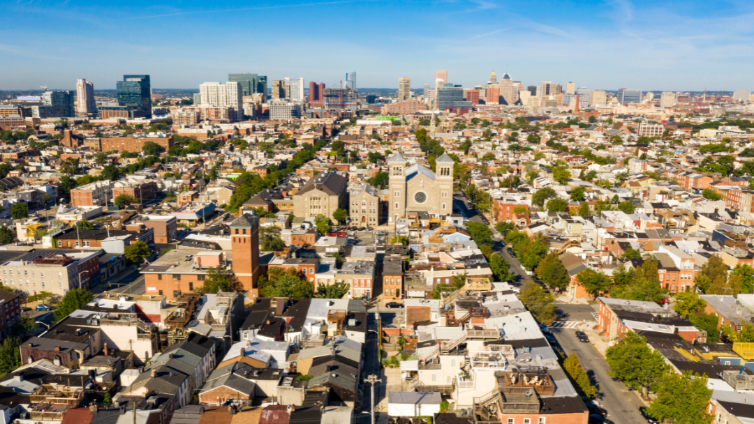 Aerial view of Baltimore's sprawling urban landscape, featuring tightly-packed residential buildings and a prominent church in the center. The city skyline is visible in the background under a clear blue sky, giving a vibrant and bustling feel.