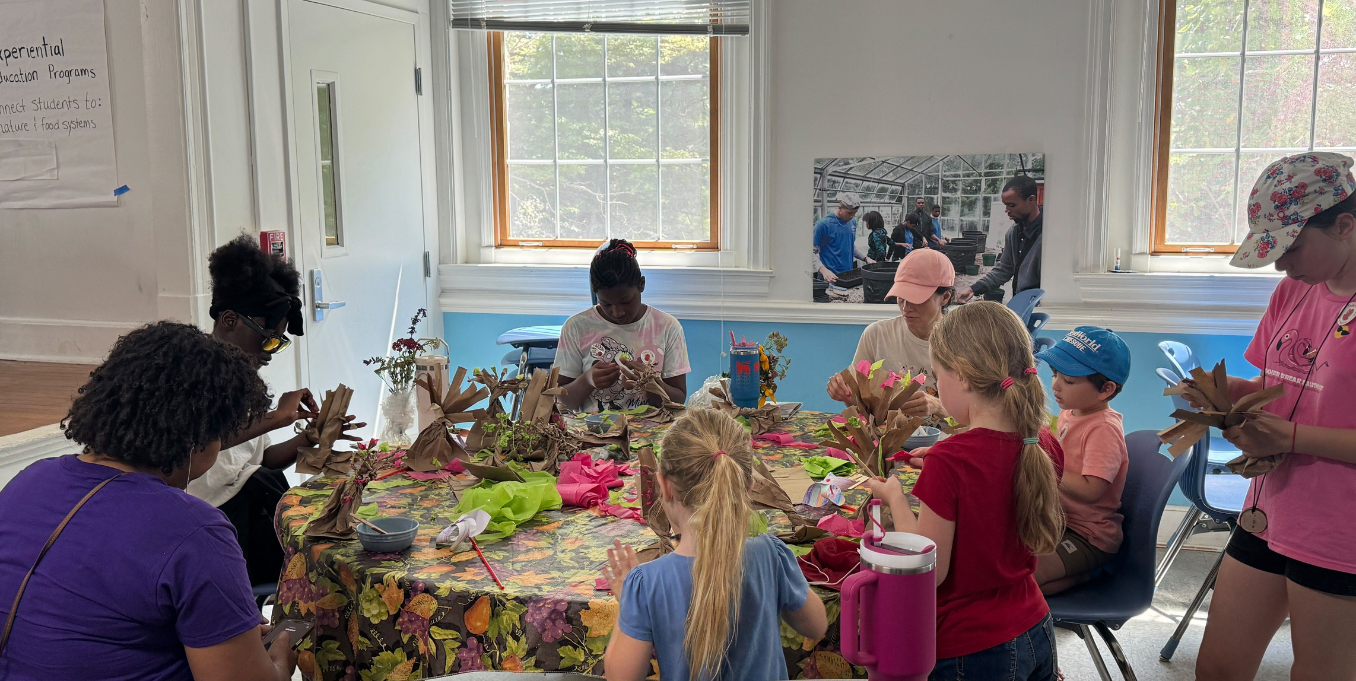A diverse group of adults and children are gathered around a table filled with craft supplies, creating art projects. The atmosphere is lively and creative.