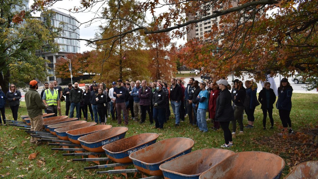 A group of people gathers outdoors for a workshop. They listen to a speaker near a line of wheelbarrows on a grassy area with autumn trees and city buildings.