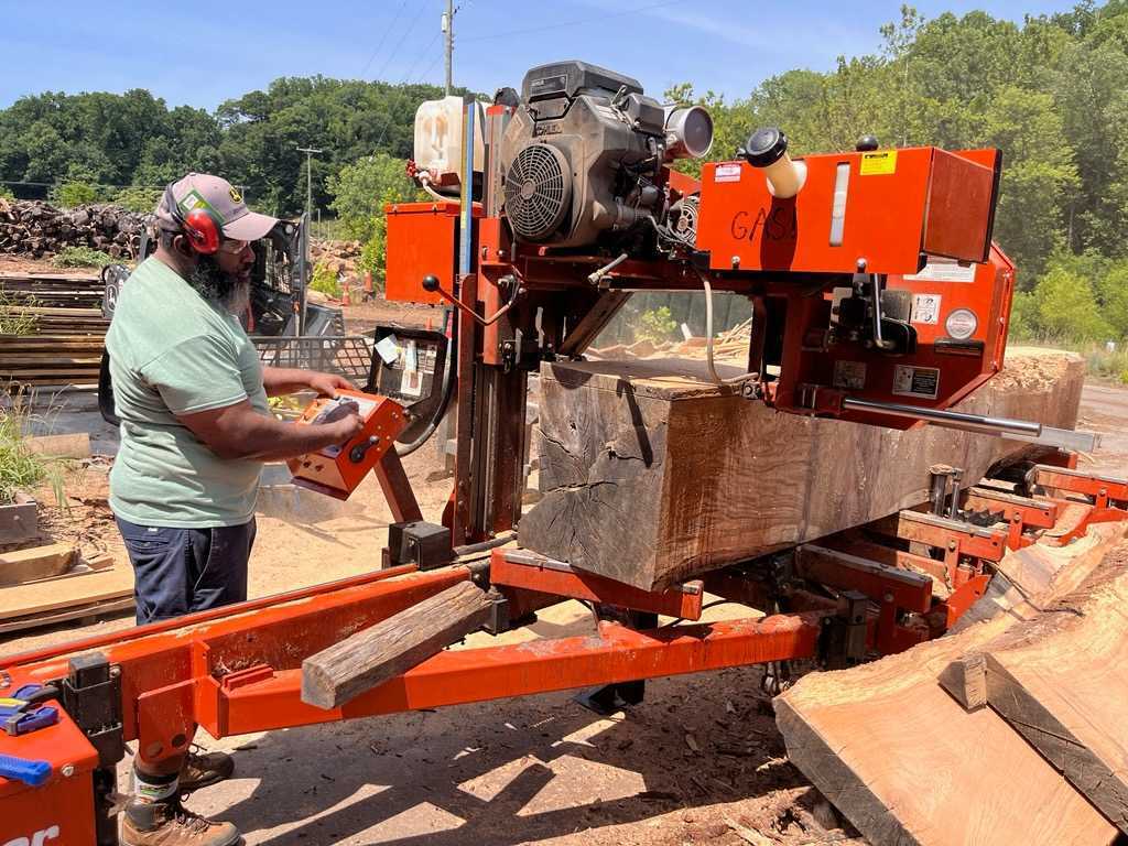 A Black man with a gray beard in a green shirt operates a red industrial sawmill, cutting a large log outdoors. He wears protective gear.